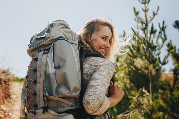 Rear view shot of smiling woman with backpack going on a camping. Caucasian female hiker on mountain looking away and smiling.